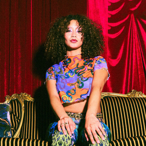 A vibrant see-through mesh crop top with a high mock neck and short sleeves. The shirt features a bold, all-over snake print pattern with colorful green, blue, yellow, and black serpents on a striking orange and purple color-block background. This Y2K-style top is a statement piece of witchy, goth, and alternative clothing, perfect for a maximalist rave outfit or edgy festival fashion from a black owned shop.